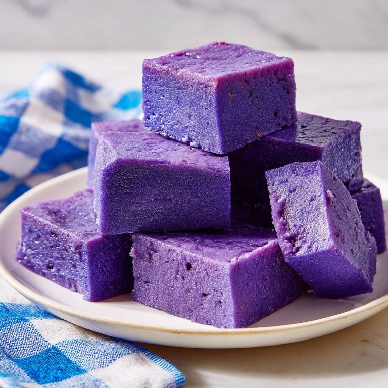 Ube Pudding Würfel gestapelt auf einem Teller mit blau-weiß kariertem Tuch daneben, perfekt cremig und schnittfest.