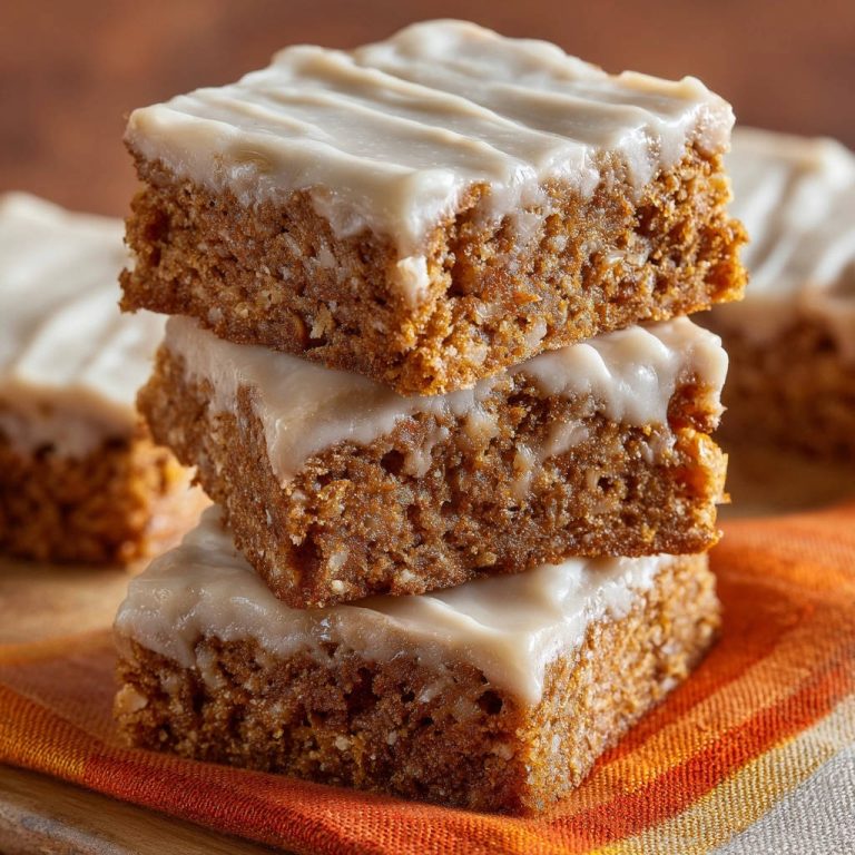 Saftige Gewürz-Bars mit cremigem Frischkäse-Topping, gestapelt auf einem orangen Tuch, Nahaufnahme des glatten Toppings.