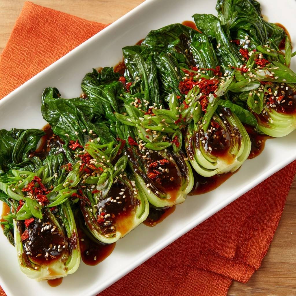 Knackiger Pak Choi mit Chili-Sesam-Soße, garniert mit Frühlingszwiebeln und Chiliflocken, serviert auf einer weißen Platte.